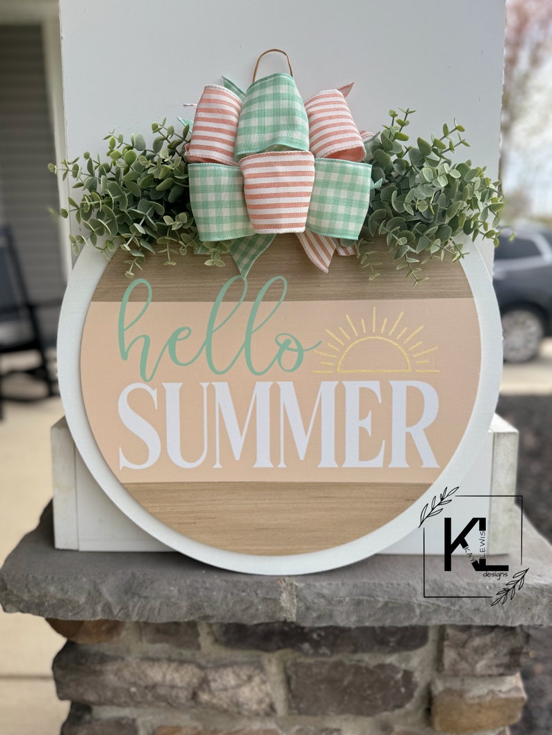 Pu&ograve; includere: Un cartello rotondo in legno con un bordo bianco, che presenta il testo "hello summer" in scrittura bianca con un grafico di sole giallo. Il cartello &egrave; decorato con un fiocco a quadri vichy verde e rosa e vegetazione.