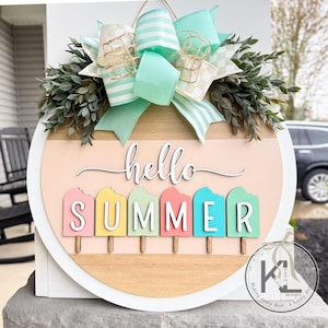 Pu&ograve; includere: Un appendiporta rotondo in legno con sfondo rosa e legno. Le parole "hello summer" sono scritte in corsivo bianco. Ci sono sei forme di ghiaccioli colorati con le lettere "SUMMER" scritte su di esse.