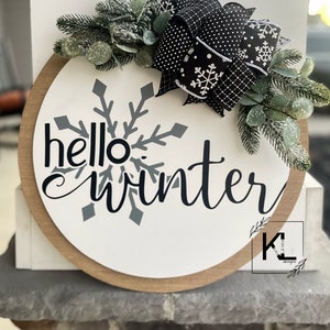 May include: A round wooden sign with the text "hello winter" in black script. A large gray snowflake is behind the text. The sign is decorated with a black and white polka dot bow and green sprigs.