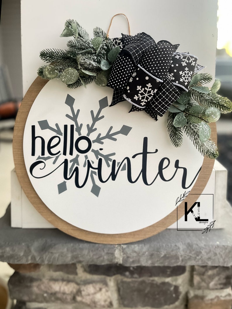 May include: A round wooden sign with the text "hello winter" in black script. A large gray snowflake is behind the text. The sign is decorated with a black and white polka dot bow and green sprigs.