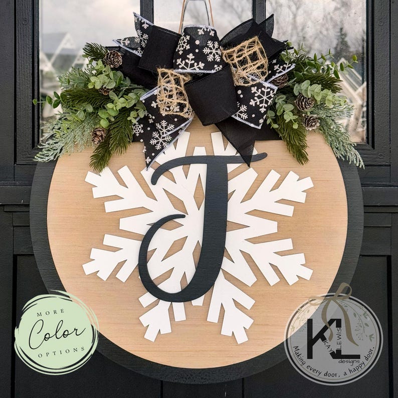 May include: A wooden round door hanger with a black border. The center features a white snowflake with a black letter "J" in the middle. The snowflake is surrounded by a burlap bow with black and white snowflake ribbon. The door hanger is decorated with greenery and pine cones.
