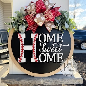 Peut inclure: Un rond de porte en bois avec un fond noir de tableau noir. Les mots "Home Sweet Home" sont &eacute;crits en blanc avec un motif de points de suture de baseball rouge autour de la lettre "H". Un n&oelig;ud rouge et blanc sur le th&egrave;me du baseball avec de la verdure verte est attach&eacute; au sommet.