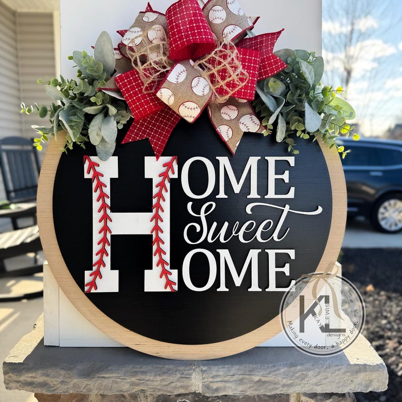 Peut inclure: Un rond de porte en bois avec un fond noir de tableau noir. Les mots "Home Sweet Home" sont &eacute;crits en blanc avec un motif de points de suture de baseball rouge autour de la lettre "H". Un n&oelig;ud rouge et blanc sur le th&egrave;me du baseball avec de la verdure verte est attach&eacute; au sommet.