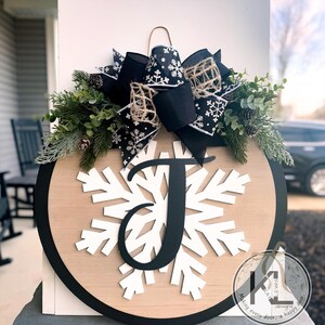 May include: A round wooden door hanger with a black border. The center features a white snowflake with a black letter "J" in the middle. A black and white bow with a burlap accent is attached to the top of the hanger.