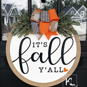 May include: A round, white door hanger with the text "It's fall y'all" in black script. A small red heart is next to the text. The door hanger is decorated with a burlap bow and orange and black checkered ribbon.