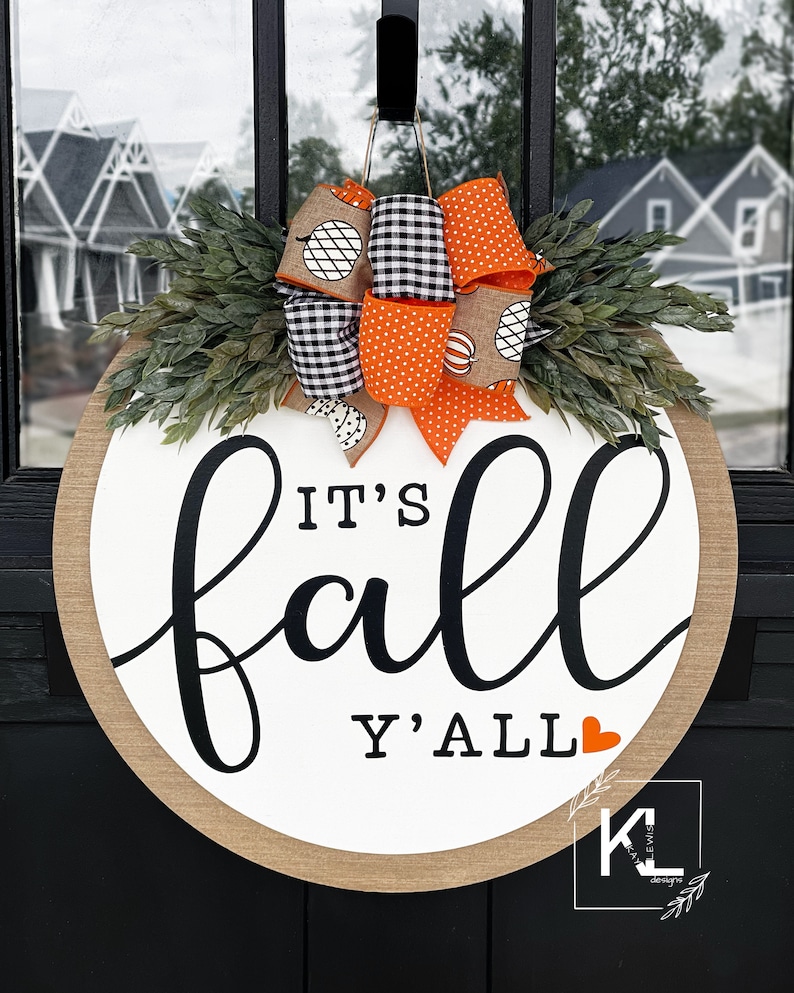 May include: A round, white door hanger with the text "It's fall y'all" in black script. A small red heart is next to the text. The door hanger is decorated with a burlap bow and orange and black checkered ribbon.
