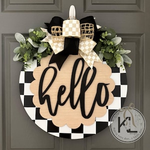 Op de afbeelding: Ronde houten deurkrans met een zwart-wit geblokte rand. De krans heeft een lichtbruin geschulpte midden met het woord "hello" in zwart schrift. Het is versierd met groen, zwart en beige geblokte strikken en een zwarte strik.