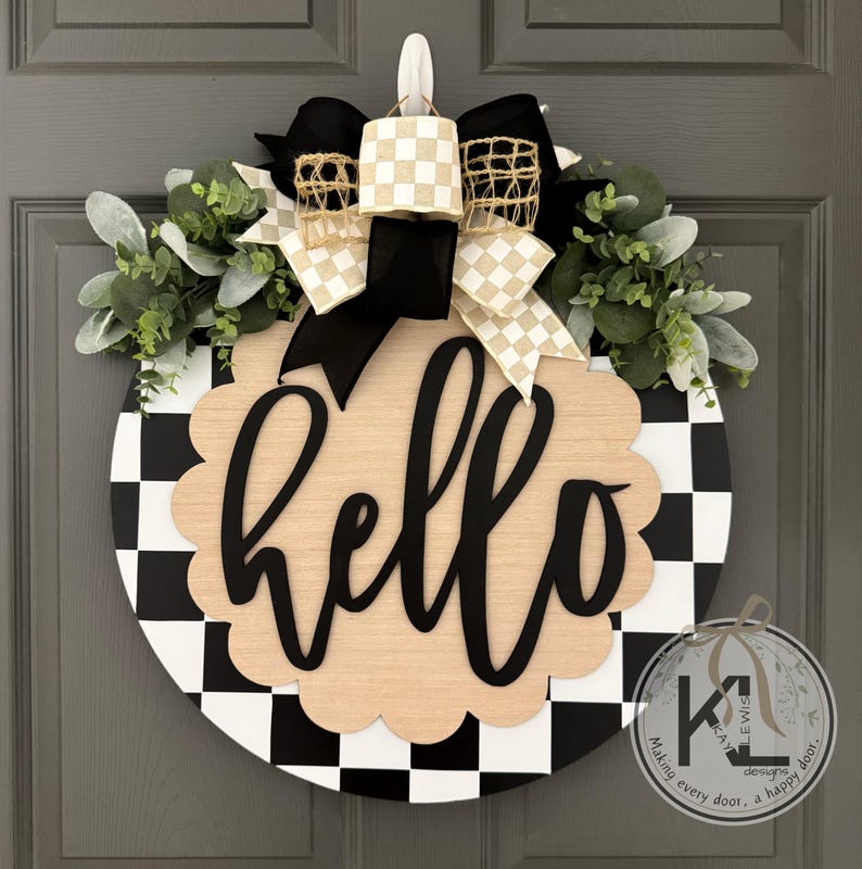 Op de afbeelding: Ronde houten deurkrans met een zwart-wit geblokte rand. De krans heeft een lichtbruin geschulpte midden met het woord "hello" in zwart schrift. Het is versierd met groen, zwart en beige geblokte strikken en een zwarte strik.