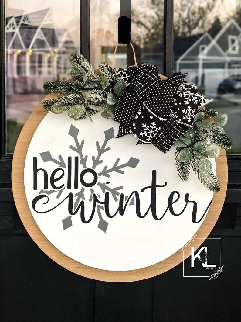 May include: A round wooden door hanger with a white background and the text "hello winter" in black cursive lettering. A gray snowflake is behind the text. A black and white polka dot bow with white snowflakes is on the top right side of the hanger. Green sprigs with white frost are around the bow.