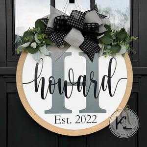 Puede incluir: Un colgador de puerta redondo de madera con fondo blanco y un lazo gris y negro. El nombre "Howard" est&aacute; escrito en letras cursivas negras con las letras "H" en gris. El texto "Est. 2022" est&aacute; escrito en negro debajo del nombre.