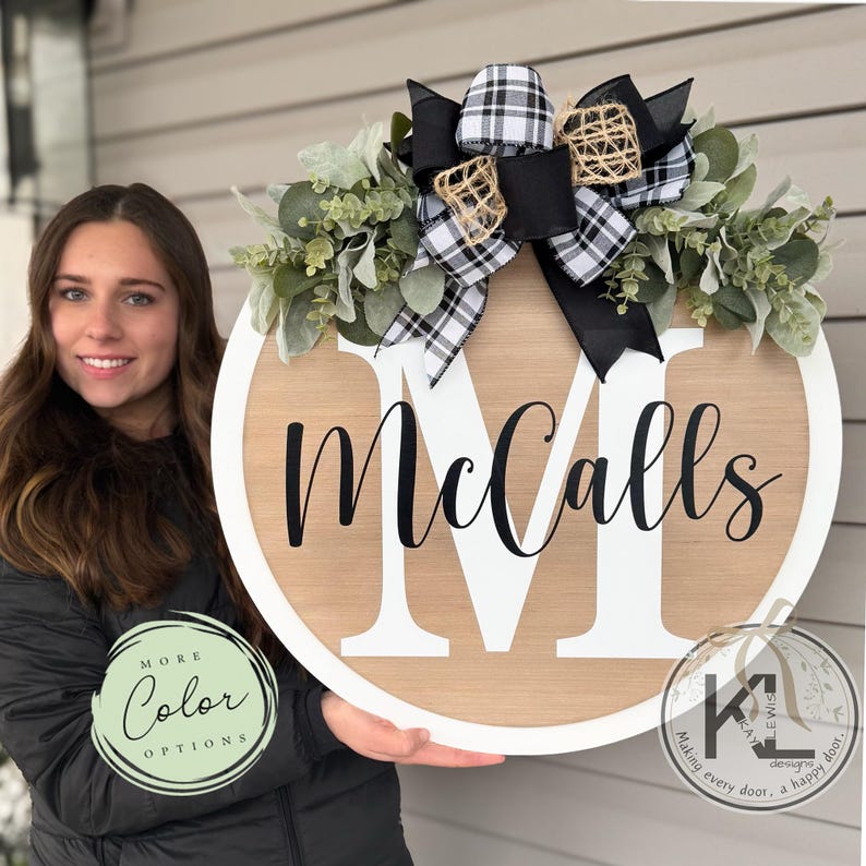 May include: A round wooden door hanger with a white border and the name "McCalls" in black script. The door hanger is decorated with a black and white plaid bow, greenery, and a burlap bow. The door hanger is held by a person wearing a black jacket.