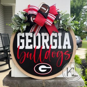 May include: A round wooden sign with the words "GEORGIA Bulldogs" in white and red lettering. The sign has a black background and a light brown border. It is decorated with a bow featuring footballs and red and white ribbons, and greenery.