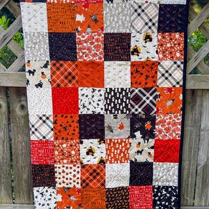 Pode incluir: Um edredom de patchwork com tema de Halloween, com quadrados de tecidos laranja, preto, branco e vermelho. O edredom apresenta v&aacute;rios padr&otilde;es, incluindo ab&oacute;boras, fantasmas, morcegos e desenhos xadrez. O edredom &eacute; contornado com um tecido preto.
