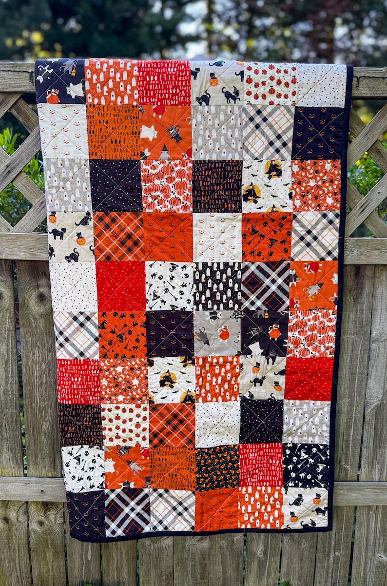 Pode incluir: Um edredom de patchwork com tema de Halloween, com quadrados de tecidos laranja, preto, branco e vermelho. O edredom apresenta v&aacute;rios padr&otilde;es, incluindo ab&oacute;boras, fantasmas, morcegos e desenhos xadrez. O edredom &eacute; contornado com um tecido preto.