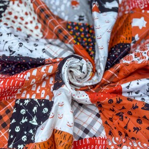 Pode incluir: Um edredom de Halloween feito &agrave; m&atilde;o com um design de patchwork. O edredom apresenta quadrados laranja, preto, branco e cinza com padr&otilde;es tem&aacute;ticos de Halloween, incluindo ab&oacute;boras, fantasmas e caveiras. O edredom &eacute; acolchoado com um padr&atilde;o de diamante.