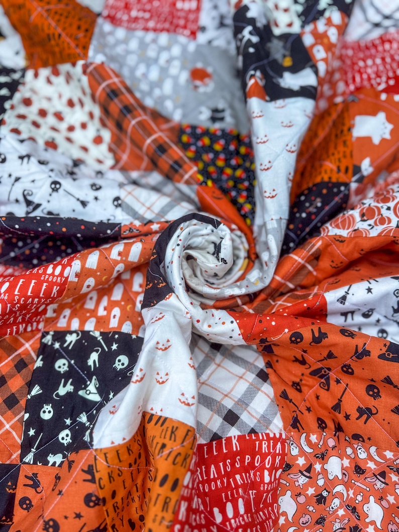Pode incluir: Um edredom de Halloween feito &agrave; m&atilde;o com um design de patchwork. O edredom apresenta quadrados laranja, preto, branco e cinza com padr&otilde;es tem&aacute;ticos de Halloween, incluindo ab&oacute;boras, fantasmas e caveiras. O edredom &eacute; acolchoado com um padr&atilde;o de diamante.