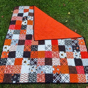 Pode incluir: Um edredom de patchwork com tema de Halloween. O edredom apresenta uma variedade de quadrados estampados em laranja, preto, branco e cinza. O verso &eacute; laranja liso. O edredom est&aacute; sobre a grama verde.
