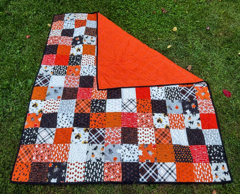 Pode incluir: Um edredom de patchwork com tema de Halloween. O edredom apresenta uma variedade de quadrados estampados em laranja, preto, branco e cinza. O verso &eacute; laranja liso. O edredom est&aacute; sobre a grama verde.