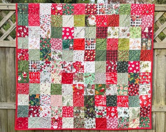 May include: A patchwork quilt with a red border, featuring a variety of Christmas-themed fabrics. The squares include patterns of Santa Claus, snowflakes, and plaid in red, green, and white. The quilt is hanging on a wooden fence.