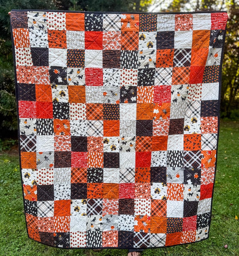 Pode incluir: Um edredom de retalhos com uma borda preta, apresentando uma variedade de tecidos com tema de Halloween. Os quadrados incluem padr&otilde;es laranja, preto, branco e cinza com morcegos, ab&oacute;boras e desenhos xadrez. O edredom &eacute; exibido ao ar livre em um gramado verde.