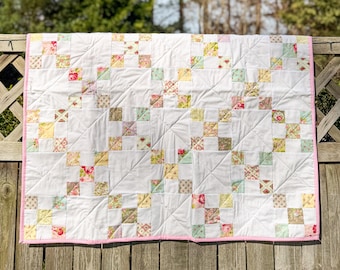 May include: A handmade quilt with a white background and a patchwork design. The quilt features squares of floral and patterned fabrics in pastel colors, including pink, yellow, and green. It has a pink border.