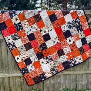 Pode incluir: Um edredom de retalhos com uma borda preta, pendurado em uma cerca de madeira. O edredom apresenta uma variedade de padr&otilde;es quadrados em laranja, vermelho, preto, branco e cinza, com motivos tem&aacute;ticos de Halloween.