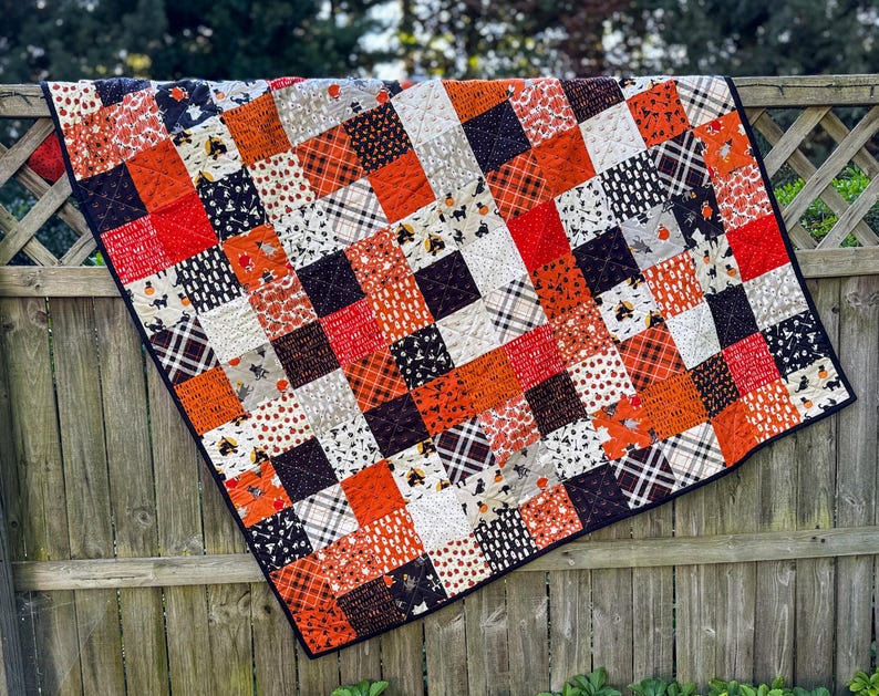 Pode incluir: Um edredom de retalhos com uma borda preta, pendurado em uma cerca de madeira. O edredom apresenta uma variedade de padr&otilde;es quadrados em laranja, vermelho, preto, branco e cinza, com motivos tem&aacute;ticos de Halloween.