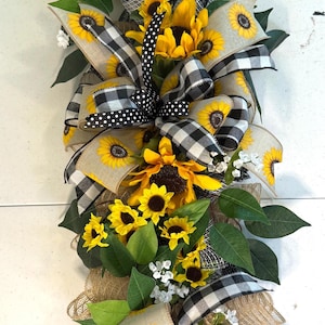 May include: A decorative floral swag featuring bright yellow sunflowers, green leaves, and white accents. The design incorporates black and white checkered and polka dot ribbons, and burlap accents. A cheerful, rustic home decor item.