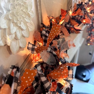May include: A close-up of a decorative garland with orange polka dots, black and white checkered, and black fabric. The garland is illuminated with small lights, creating a warm glow. A white floral wall decoration is visible in the background.