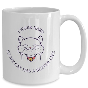 Può includere: Tazza in ceramica bianca con l'illustrazione di un gatto sorridente e la scritta "I WORK HARD SO MY CAT HAS A BETTER LIFE" in viola scuro. Il gatto ha un collare rosso con un campanello dorato. La tazza ha un manico grande e curvo.