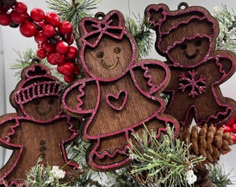 May include: Three wooden gingerbread people ornaments with pink glitter accents. The ornaments are shaped like a girl, a boy, and a gingerbread man. The ornaments are hanging on a green pine branch with red berries.
