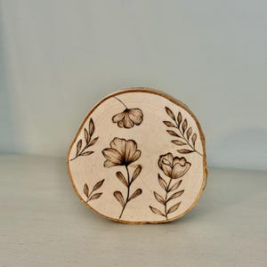 May include: A round, natural wood slice with a light brown hue. The wood slice features a floral design, with three flowers and several leaves, all in a dark brown color. The wood slice is a decorative item.