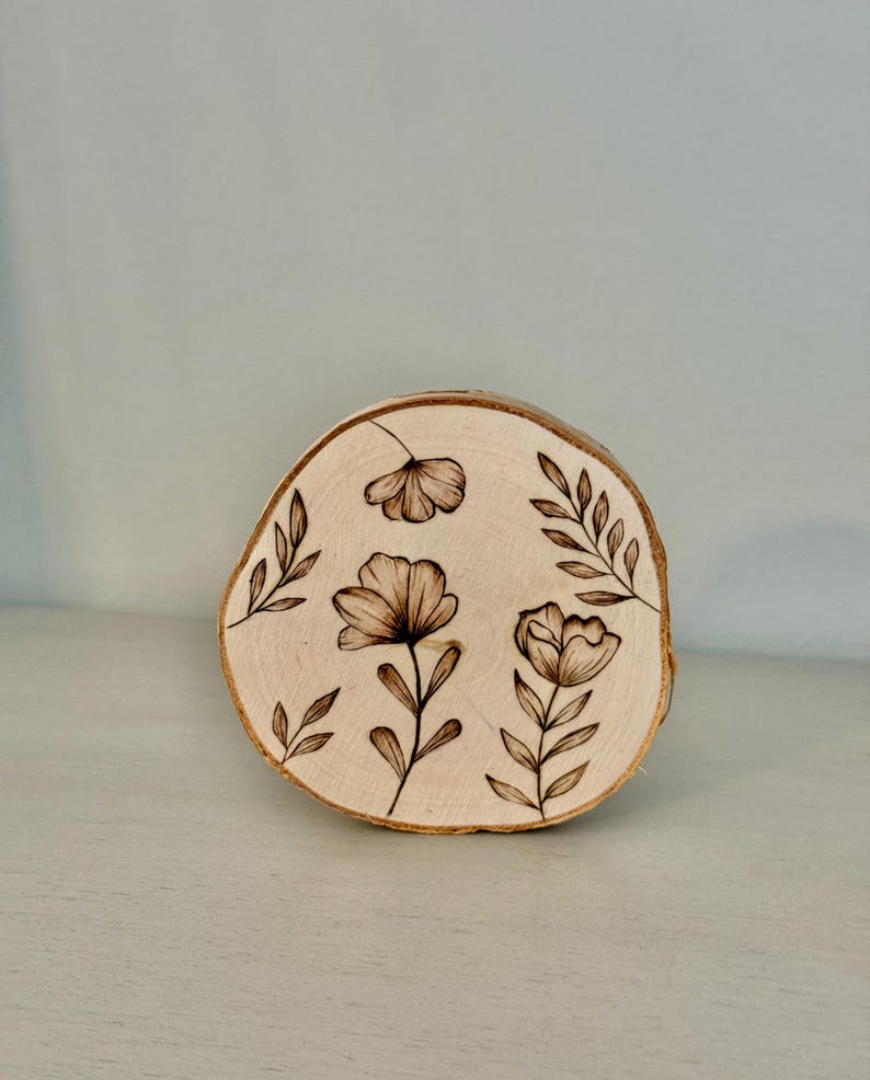 May include: A round, natural wood slice with a light brown hue. The wood slice features a floral design, with three flowers and several leaves, all in a dark brown color. The wood slice is a decorative item.