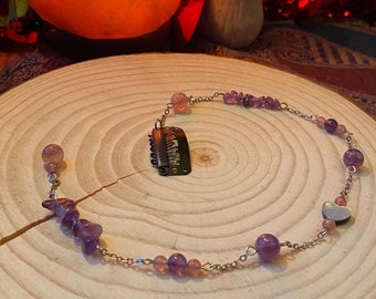 May include: A silver chain necklace with purple and pink beads, a silver heart charm, and a silver hair clip. The necklace is on a wooden surface.