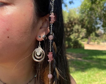May include: A close-up of a person's hair, styled in a braid, adorned with silver hair rings and a chain of pink gemstones. The person is also wearing a silver spiral earring and a delicate necklace.