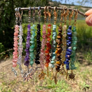 May include: A collection of ten beaded keychains featuring various polished gemstone chips in shades of pink, purple, green, black, red, orange, brown, yellow, and blue, each attached to a gold or silver metal lobster clasp and a charm.