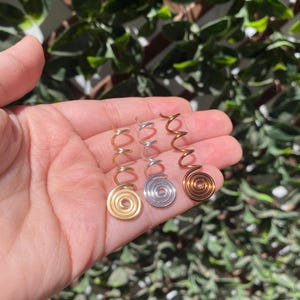 May include: Three spiral hair accessories in gold, silver, and copper tones. Each accessory features a coiled spiral design at the base and a curved, elongated top. The accessories are held in a hand against a backdrop of green foliage.