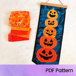 May include: A vertical fabric wall hanging featuring a stack of five orange jack-o'-lanterns on a dark blue marbled background, suspended from a wooden rod. Beside it lies a stack of orange and red patterned fabric squares and metal scissors.