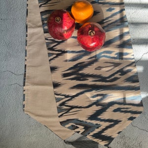 May include: A beige and blue patterned table runner with tassels on the ends. Three fruits are on top of the runner: two red pomegranates and one orange.
