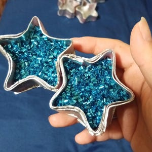 May include: Two silver star-shaped containers filled with small, shiny, turquoise beads. The containers are held in a hand, with another star-shaped container visible in the background. The beads fill the star shapes, creating a textured surface.