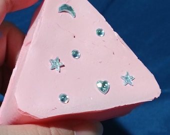 May include: A triangular-shaped pink object with a smooth matte surface, decorated with several small, light blue, reflective, gem-like embellishments in the shapes of a crescent moon, stars, a heart, and small circles scattered across the front face.