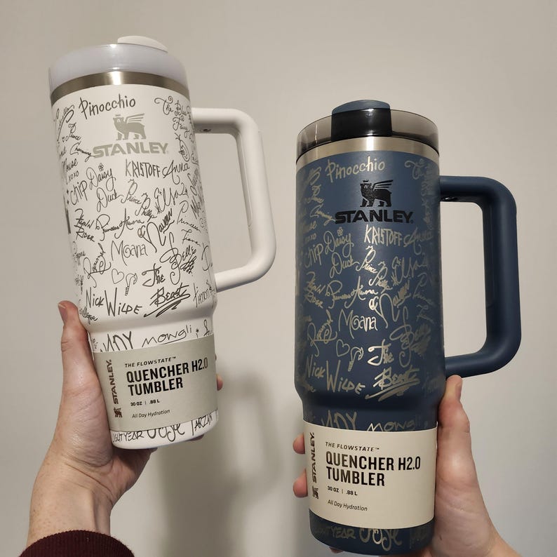 May include: Two stainless steel travel tumblers with a white and blue finish. Both tumblers feature a silver inscription with the Stanley logo and the text "The Flowstate Quencher H2.0 Tumbler 30 oz | 887 ml All Day Hydration".