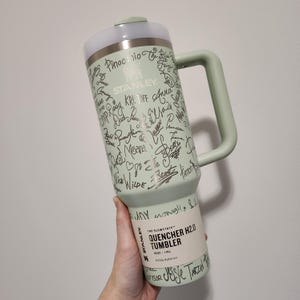 May include: A light green Stanley Quencher H2.0 Tumbler with a silver lid and handle. The tumbler is covered in silver signatures. The text "The Flowstate" and "40 oz | 1.18 L" are printed on the tumbler.