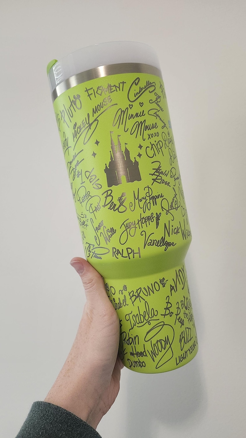 May include: A green stainless steel tumbler with a silver lid. The tumbler is covered in silver writing, including the names of Disney characters like Mickey Mouse, Cinderella, and Bruno. The tumbler also features a silver silhouette of Cinderella's castle.
