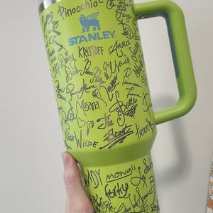 May include: A green Stanley travel mug with a black handle. The mug is covered in black marker drawings of Disney characters and their names, including Pinocchio, Kristoff, Moana, Belle, and Mowgli.