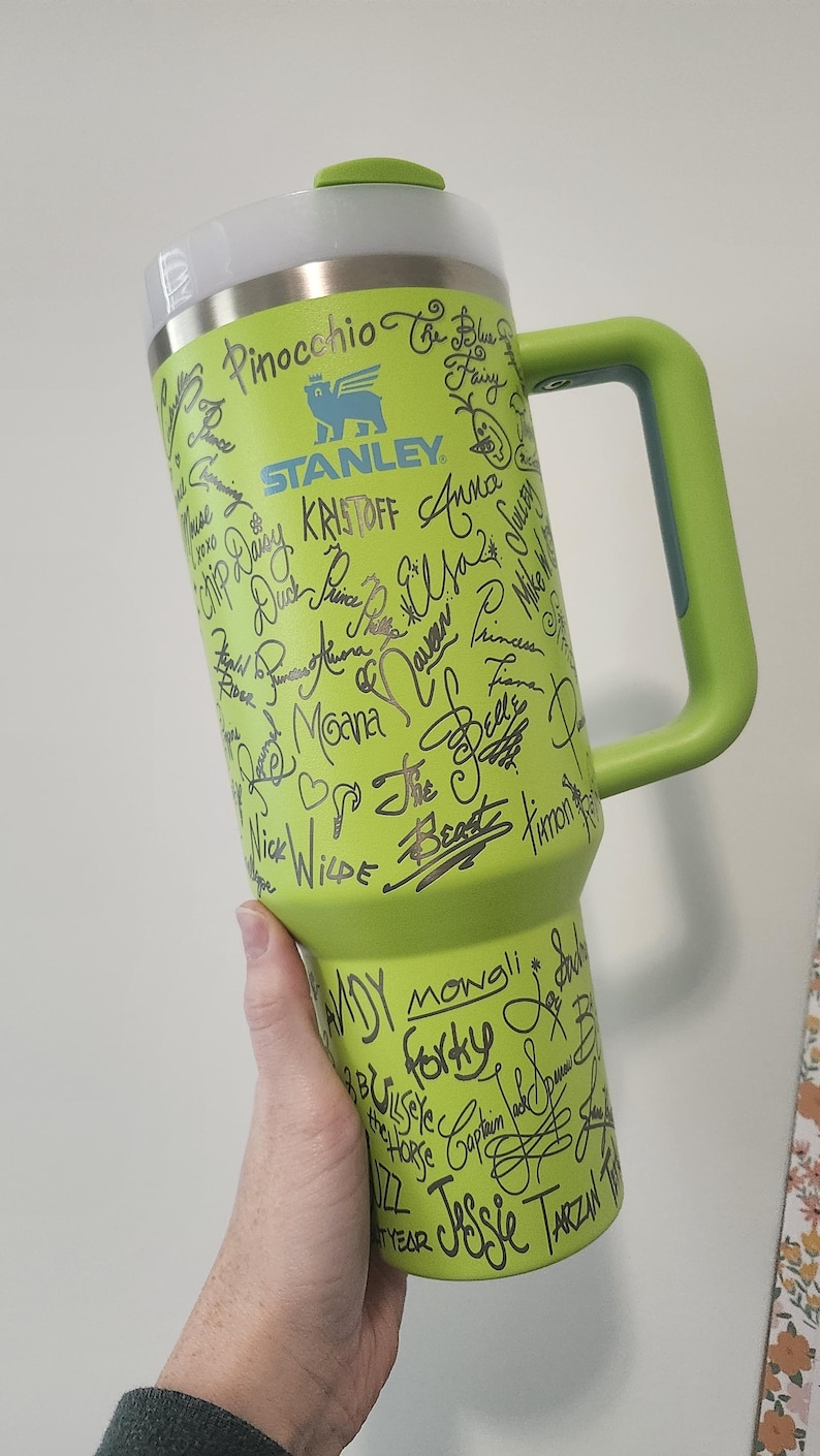 May include: A green Stanley travel mug with a black handle. The mug is covered in black marker drawings of Disney characters and their names, including Pinocchio, Kristoff, Moana, Belle, and Mowgli.