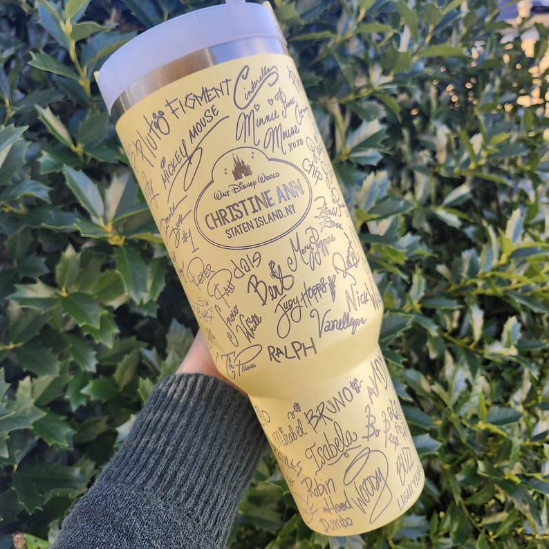May include: A yellow insulated tumbler featuring signatures and the text "CHRISTINE ANN STATEN ISLAND, NY" with a Disney World logo. The tumbler includes signatures of Disney characters like Mickey Mouse and Cinderella.