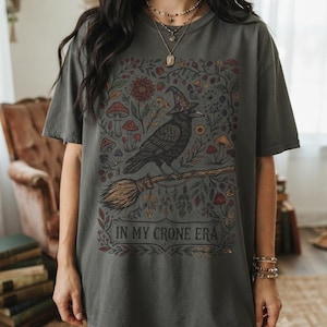 May include: A charcoal gray t-shirt featuring a graphic print of a black crow wearing a witch hat, perched on a broomstick. The design includes colorful mushrooms, flowers, and foliage, with the text "IN MY CRONE ERA" printed at the bottom.