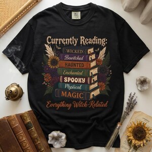 May include: A black t-shirt featuring a graphic of a stack of seven books with the text Currently Reading: and Everything Witch-Related. The book spines are labeled Wicked, Bewitched, Haunted, Enchanted, Spooky, Mystical, and Magic, surrounded by flowers.