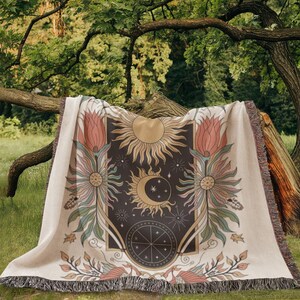 Peut inclure: Une couverture en tissage de style tapisserie avec un fond beige et un motif brun foncé, noir et or. Le motif représente un soleil, une lune et des étoiles avec des accents floraux.